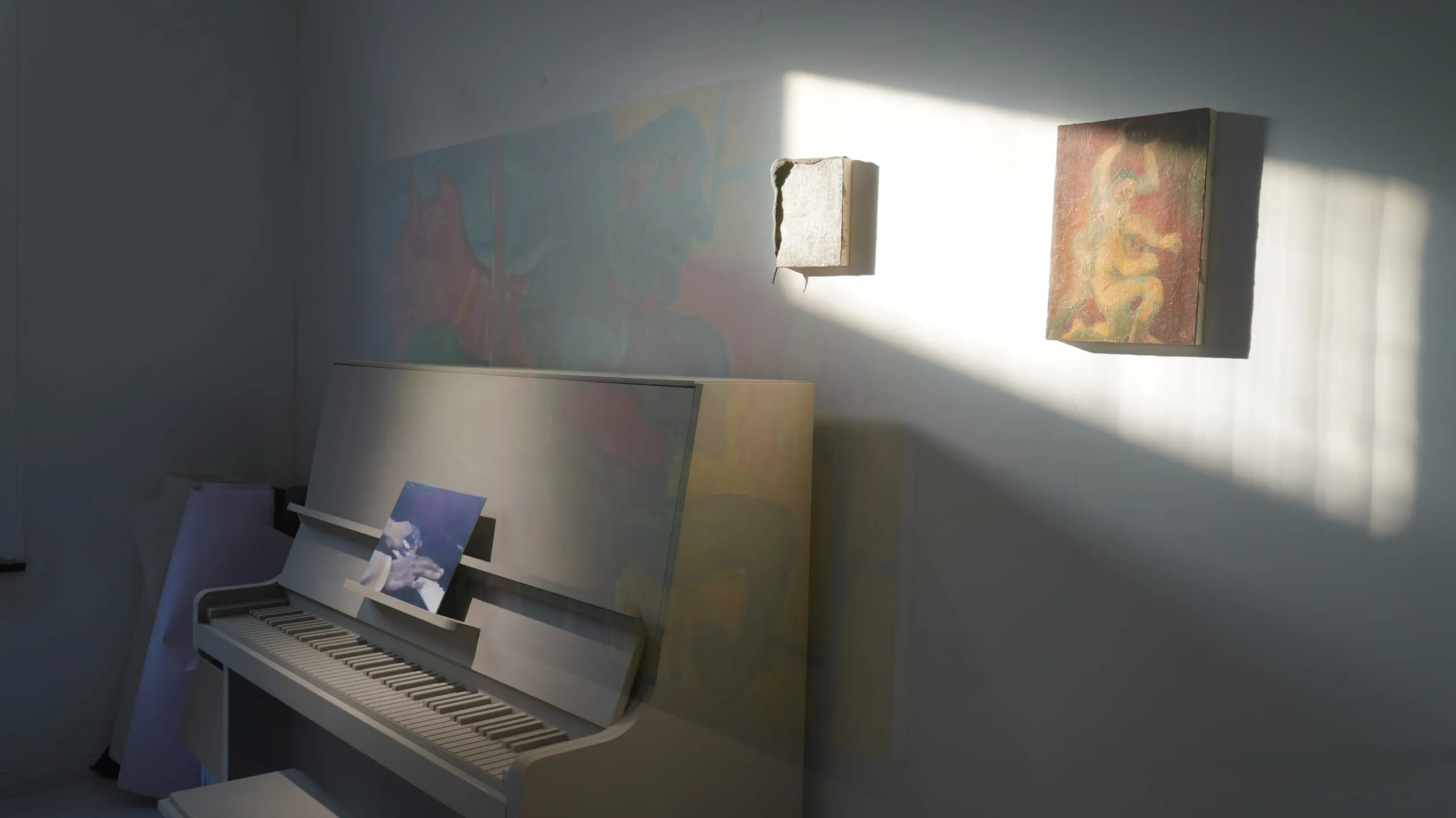 Daylight crossing a quiet corner of the space, an upright piano and two small framed works.